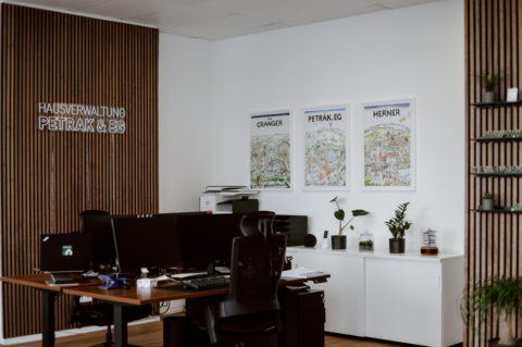 Zwei Schreibtische im Büro der Hausverwaltung Petrak & EG. Sie sind mit zwei Bildschirmen ausgestattet. An der Wand hängen Bilder und das Logo.
