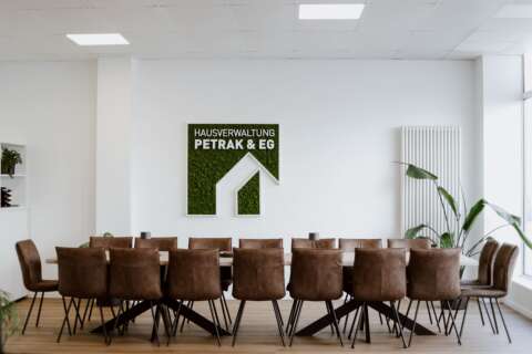 Das Besprechungszimmer der Hausverwaltung, ein großer Tisch mit vielen braunen Stühlen, im Hintergrund sowie das Firmenlogo in Moosoptik an der Wand.
