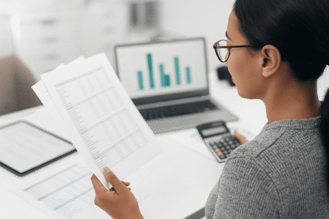 Eine Frau an einem Schreibtisch hält Dokumente in der Hand und rechnet etwas aus.