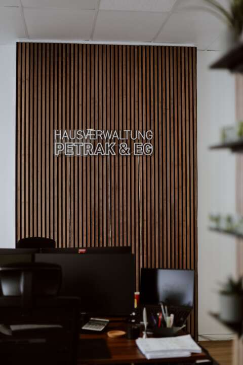 Zwei Arbeitsplätze mit Bildschirm, Bürostuhl und weiteren Utensilien, im Hintergrund Hausverwaltung Petrak & EG als Leuchtschrift an der Wand.